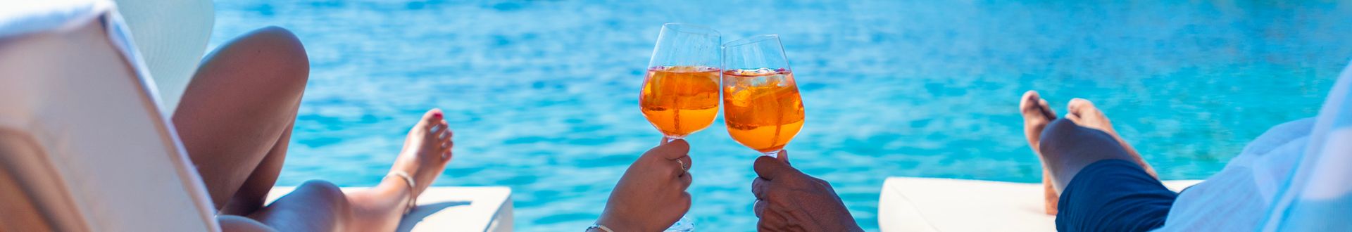 Zwei Personen stoßen mit gekühlten Getränken über dem türkisblauen Meer an, während sie entspannt auf einer Sonnenliege sitzen