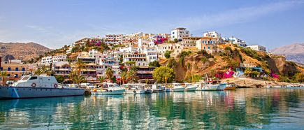Blick auf die Altstadt von Heraklion auf Kreta mit weißen Häusern, bunten Blumen und Booten im Hafen vor felsigem Ufer.