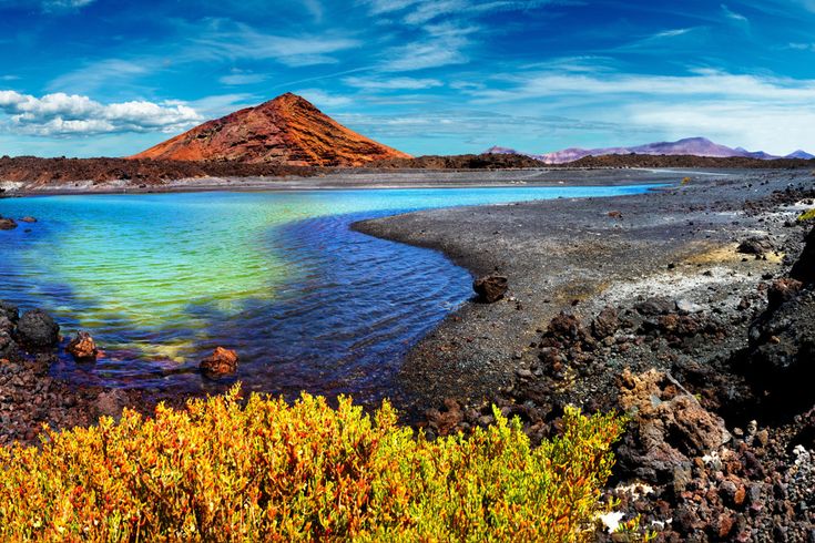 Landschaft mit blau-grünem See, umgeben von schwarzer Vulkanlandschaft und einem roten Vulkan im Hintergrund