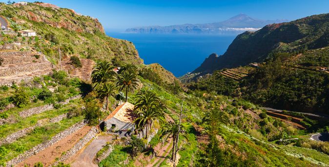Berglandschaft auf La Gomera mit terrassierten Feldern, Palmen und Blick auf den Atlantik und entfernten Vulkan