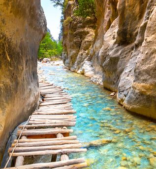 Ein schmaler Wasserlauf mit klarem, blauem Wasser fließt durch eine enge Schlucht, umgeben von hohen, steilen Felsen. Ein Holzsteg führt über das Wasser.