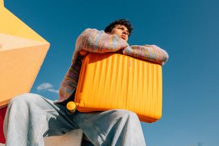 Junge mit farbenfrohem Pullover sitzt im Freien und lehnt sich an einen orangefarbenen American Tourister Koffer vor klarem blauem Himmel.