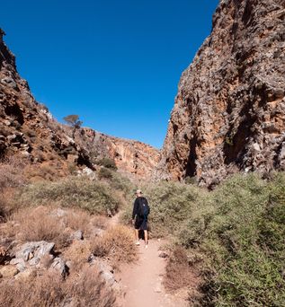 Wanderer mit schwarzem Rucksack auf schmalem Pfad zwischen hohen, felsigen Schluchtwänden unter blauem Himmel.