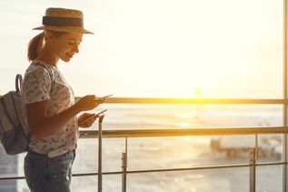 Frau mit Strohhut, Rucksack und T-Shirt steht am Flughafenfenster und hält ein Smartphone, Sonnenuntergang im Hintergrund.