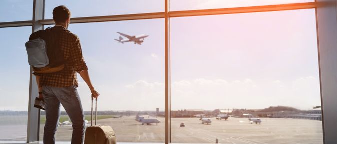 Reisender mit Rucksack und Rollkoffer blickt durch die Fenster eines Flughafens auf ein startendes Flugzeug