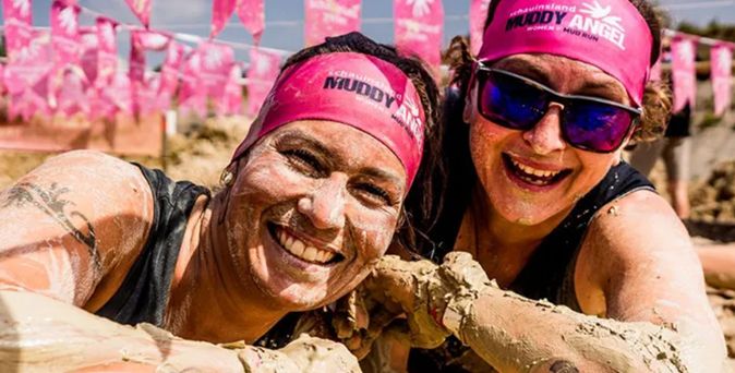 Zwei Teilnehmende beim Muddy Angel Run, mit Schlamm bedeckt an einem Hindernis, tragen pinke Stirnbänder mit Event-Branding und posieren vor Fahnen der Veranstaltung