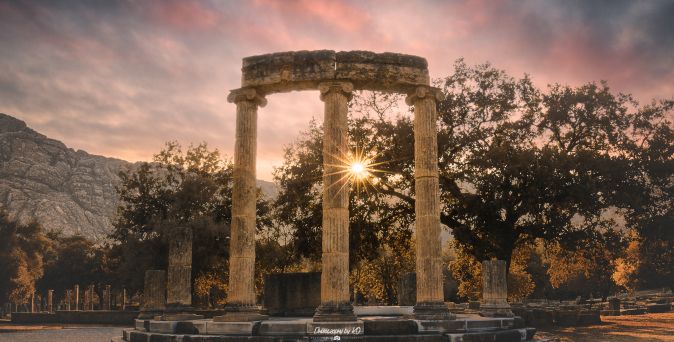 Sonnenuntergang hinter den antiken Säulen des Tempels von Olympia, umgeben von Bäumen und Bergen.