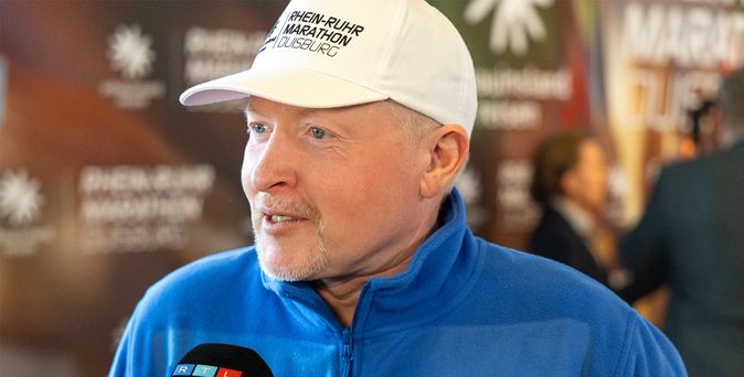 Joey Kelly trägt eine weiße Kappe und eine blaue Jacke mit dem Schriftzug 'RHEIN-RUHR MARATHON DUISBURG' und wird von einem Mikrofon mit den Logos von RTL und ntv interviewt.