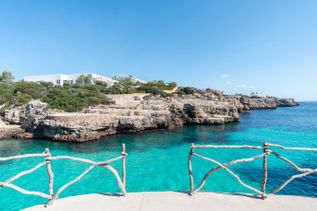 An der Bucht Cala en Brut auf Menorca leuchtet das türkisfarbene Meer vor steilen Felsen und weißen Häusern unter blauem Himmel