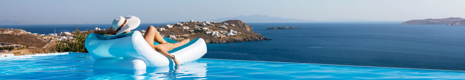 Frau liegt auf der Luftmatratze im Infinity-Pool und hat einen Sonnenhut auf. Der Blick geht aufs dunkelblaue Meer und eine Insel am Horizont.