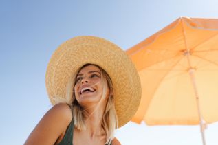 Frau mit großem, geflochtenem Strohhut sitzt unter einem orangefarbenen Sonnenschirm an einem sonnigen Tag