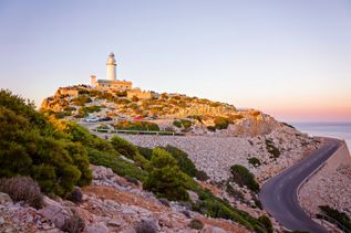 Leuchtturm auf einem Felsen am Cap de Formentor auf Mallorca, umgeben von Felslandschaft, Vegetation und einer kurvigen Küstenstraße, aufgenommen bei Sonnenuntergang