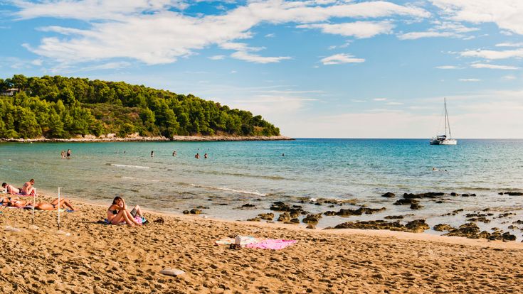 Badegäste am Sandstrand Vela Pržina in Kroatien mit flachem, klarem Meerwasser, bewaldeter Küste und einem Segelboot vor der Küste