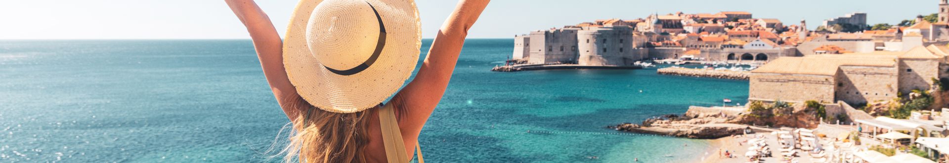Person mit Sonnenhut und Rucksack blickt auf die Altstadt von Dubrovnik in Kroatien mit Stadtmauer, türkisblauer Adria, Küstenpromenade und Strand im Vordergrund