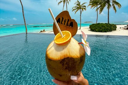 Hand hält eine Kokosnuss mit Trinkhalm, Orangenscheibe, Blüte und Holzschild mit der Aufschrift 'Alila' vor einem Pool und Strand mit Palmen