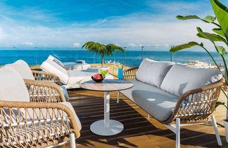 Großer runder Hotelpool mit Liegestühlen und Palmen, angrenzendes Hotelgebäude links, Hügel mit Häusern und Meer im Hintergrund unter blauem Himmel.