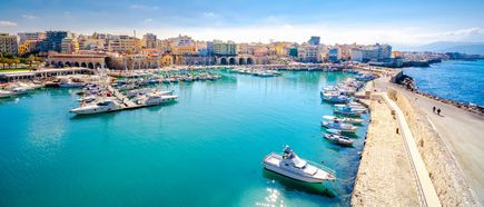 Hafen von Heraklion auf Kreta mit mehreren Booten im Wasser und einer Ufermauer, im Hintergrund Stadtgebäude unter klarem Himmel.