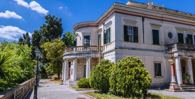 Neoklassizistisches Gebäude mit Säulen und Balkon umgeben von Bäumen und Sträuchern unter blauem Himmel
