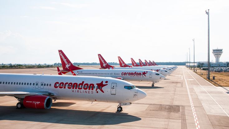 Corendon-Airlines-Flotte mit mehreren Boeing-737-800-Flugzeugen, aufgereiht auf dem Vorfeld eines Flughafens, rote Leitwerke mit Corendon-Logo, Flugverkehrskontrollturm im Hintergrund