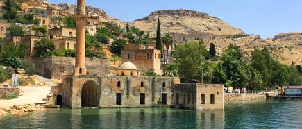 Historisches Steingebäude mit Minarett am Flussufer in der Region Gaziantep, teilweise vom Wasser umgeben, mit felsigen Hügeln im Hintergrund