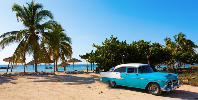 Blauer Oldtimer-Pkw auf sandigem Strand mit Palmen und Meer im Hintergrund auf Kuba