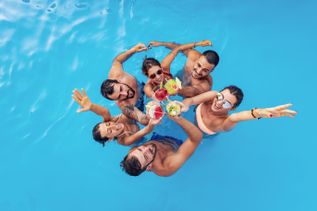Gruppe junger Menschen im Pool stößt mit bunten Cocktails an und lacht in die Kamera