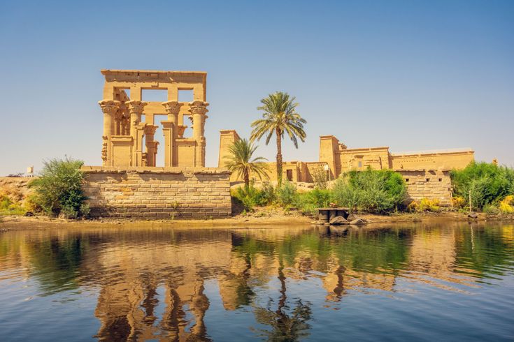 ae-Tempel in Ägypten mit antiken Steinsäulen am Ufer des Nils, Spiegelung im Wasser, Palmen und weitere Tempelbauten vor blauem Himmel