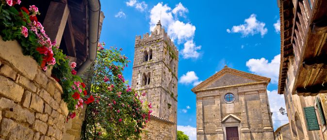 Kirche Maria Himmelfahrt in Hum, Kroatien, mit historischem Steinturm und blühenden Blumen vor blauem Himmel