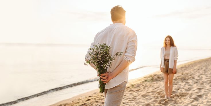 Mann hält einen Blumenstrauß hinter dem Rücken und geht eine Strandpromenade entlang auf eine Frau zu; warmes Licht über dem Wasser