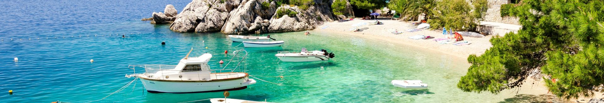 Bucht an der Makarska Riviera in Brela, Kroatien, mit kristallklarem türkisfarbenem Wasser, mehreren vor Anker liegenden Booten, hellem Kiesstrand und von Pinien sowie Felsen gesäumter Adriaküste