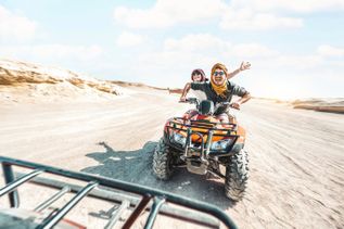 Zwei Personen fahren fröhlich auf einem Quad auf einem sandigen Weg