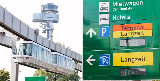 Schwebebahn vor dem Tower des Düsseldorfer Flughafens neben einem grünen Verkehrsschild mit Hinweisen zu Mietwagen, Hotels und Langzeitparkplätzen am Terminal