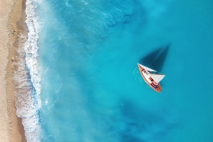 Luftaufnahme eines Segelboots auf türkisblauem Meer nahe eines Sandstrandes.