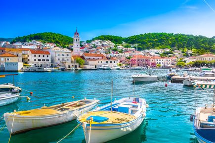 Kleine Boote im Hafen von Pučišća vor einer Küstenstadt mit Natursteinhäusern, Kirchturm und grünen Hügeln auf der Insel Brač