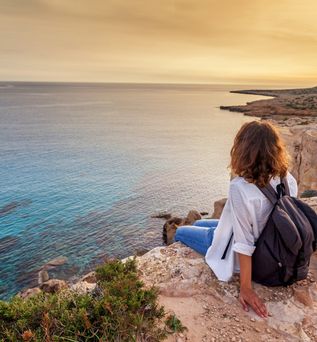 Frau mit schwarzem Rucksack sitzt auf Felsen und blickt auf das ruhige Meer bei Sonnenuntergang an der Küste von Zypern