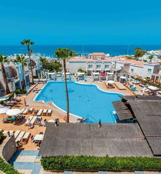Großer Hotelpool mit mehreren Sonnenliegen und Sonnenschirmen, umgeben von weißen Gebäuden und Palmen, im Hintergrund das Meer.