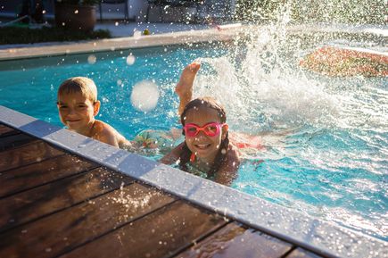 Zwei Kinder schwimmen in einem Pool mit spritzendem Wasser am Beckenrand, während Sonnenlicht auf die Wasseroberfläche fällt