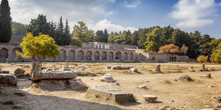 Ruinen des antiken Asklepieions auf Kos mit einer langen Arkadenreihe und verstreuten Steinen auf trockenem Boden vor bewaldetem Hintergrund
