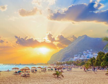 Strand mit Sonnenschirmen und Palmen bei Sonnenuntergang, im Hintergrund Berge und Häuser am Hang.