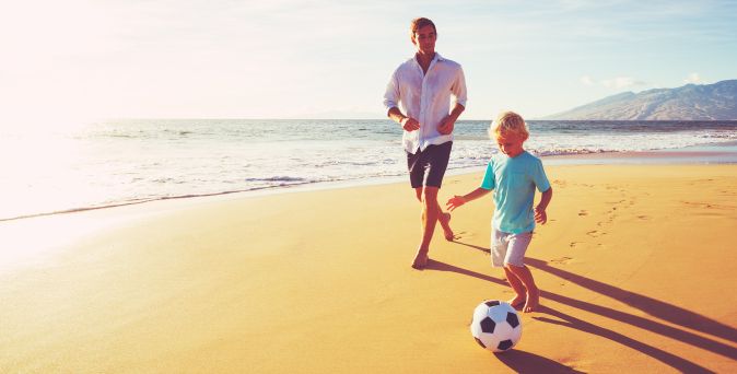 Vater und Sohn spielen barfuß am Strand Fußball bei klarem Himmel und beginnendem Sonnenuntergang.
