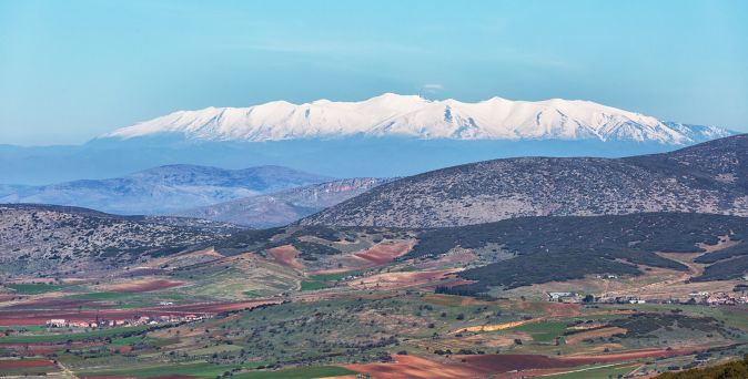 Schneebedeckter Berggipfel im Hintergrund, davor hügelige Landschaft mit grünen und braunen Feldern unter blauem Himmel.
