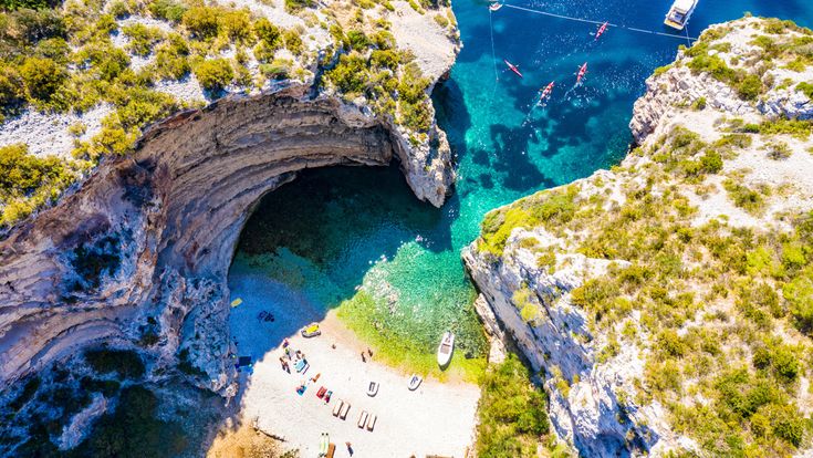Vogelperspektive auf die Stiniva-Bucht in Kroatien mit steilen Felswänden, kleinem Kiesstrand, türkisfarbenem Meer und mehreren Booten vor der Küste
