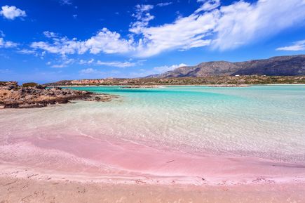 Kretas flacher Strand mit rosa schimmerndem Sand trifft auf klares, türkisfarbenes Wasser. Dahinter liegen Felsen und eine bergige Landschaft unter einem blau-weißen Himmel.