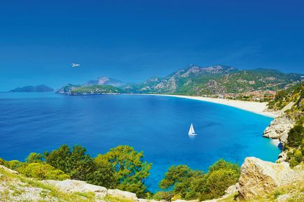 Blick auf den Ölüdeniz-Strand in Fethiye, Türkei, mit türkisblauem Meer, weißem Sandstrand, grünen Bergen und einem Segelboot unter klarem Himmel