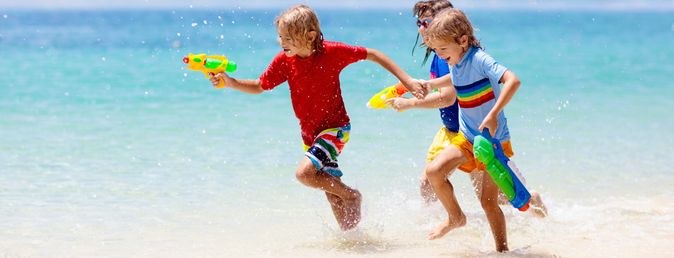 Kinder rennen lachend am Strand entlang und spritzen mit Wasserpistolen, während sie durch das flache Meerwasser spielen
