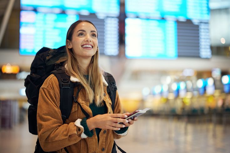 Junge Frau mit Rucksack, Smartphone und Reisepass lächelt in einer Flughafenhalle vor den großen Anzeigetafeln