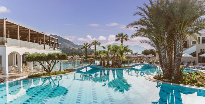 Hotelpool mit Palmen und Liegestühlen vor weißem Gebäude und Bergen im Hintergrund