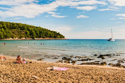 Sandstrand Vela Pržina in Kroatien mit Badegästen im flachen Meer, ruhiger Bucht, grüner Küstenlandschaft und Segelboot am Horizont