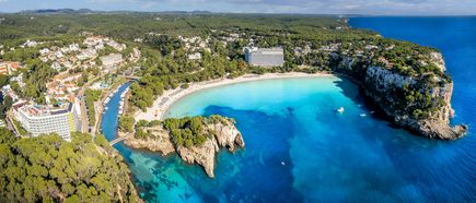 Luftaufnahme der Bucht Cala Galdana auf Menorca mit türkisblauem Wasser, Sandstrand mit Sonnenschirmen, Hotels und umliegendem Pinienwald