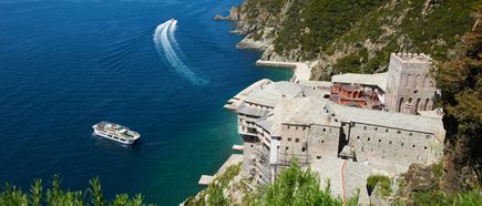 Blick von oben auf ein großes Klostergebäude an einer steilen Küste, davor ein Ausflugsboot auf blauem Wasser mit sichtbarer Bugwelle.